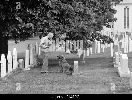 ELDERLY MAN HAT OVER HIS HEART AND PET DOG VISITING GRAVE OF LOVED ONE IN CHURCHYARD CEMETERY Stock Photo