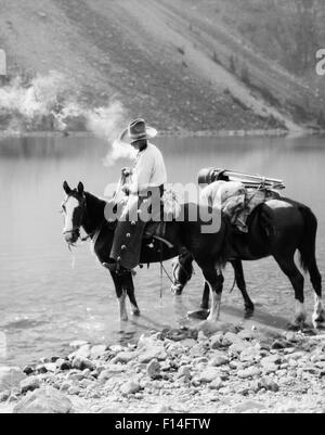 1920s 1930s MAN COWBOY ON HORSE WITH PACK HORSE BY MORAINE LAKE ALBERTA CANADA Stock Photo