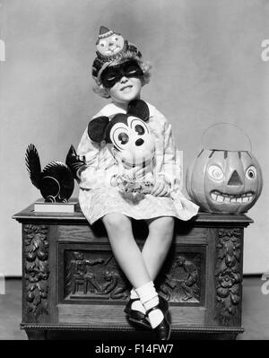 1930s LITTLE GIRL HOLDING MICKEY MOUSE TOY WEARING MASK CLOWN HAT LOOKING AT CAMERA SITTING BESIDE BLACK CAT HALLOWEEN PUMPKIN Stock Photo