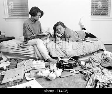 1980s TWO TEENAGE AFRICAN AMERICAN GIRLS IN CLUTTERED BEDROOM ONE TALKING ON THE PHONE ONE READING MAGAZINE Stock Photo