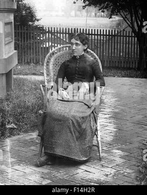 1890s 1900s TURN OF THE 20TH CENTURY PORTRAIT SEVERE WOMAN SITTING IN WICKER CHAIR IN BACKYARD BY HOUSE Stock Photo