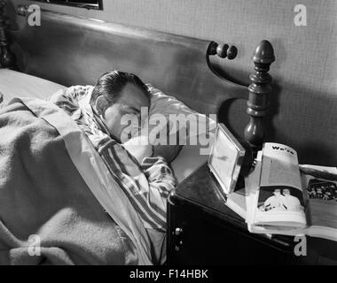 1950s MIDDLE AGED MAN SLEEPING ASLEEP STRIPED PAJAMAS CLUTCHING A PILLOW NIGHT TABLE ALARM CLOCK MAGAZINE Stock Photo