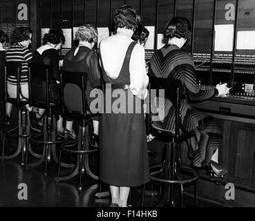 1930s BACK VIEW OF SEATED WOMEN SWITCHBOARD OPERATORS AND STANDING SUPERVISOR Stock Photo