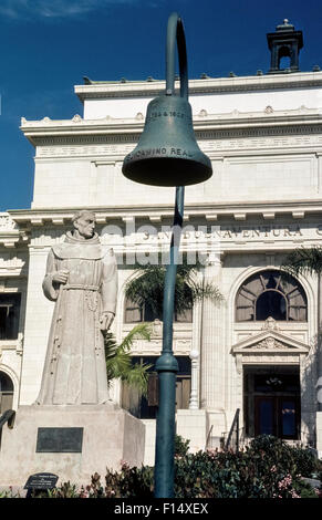 A cast iron bell on El Camino Real (The Royal Road) of 21 Spanish missions stand stands next to a concrete statue of mission founder Father Junipero Serra at the San Buenaventura city hall in Ventura, California, USA. Sculpted in 1936 by John Palo-Kangas, the nine-foot tall monument deteriorated over time and was replaced by a bronze copy in 1989. The two dates inscribed on the bell denote the founding of the first mission in San Diego in 1769 and the dedication in 1906 of the first of several hundred identical bells marking the mission trail up and down the state. Stock Photo