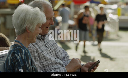 Happy couple using cell phone on bench at amusement park fun fair Stock Photo