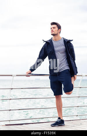 Portrait of a handsome sportsman stretching legs outdoors in the morning with sea on background Stock Photo