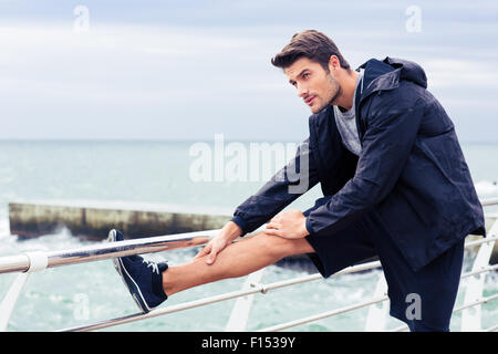 Portrait of a handsome sportsman stretching legs outdoors in the morning with sea on background Stock Photo