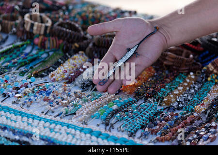 A man holds a custom beaded bracelet shaped as a lizard over a table of handmade jewelry at a beach market in Belize. Stock Photo