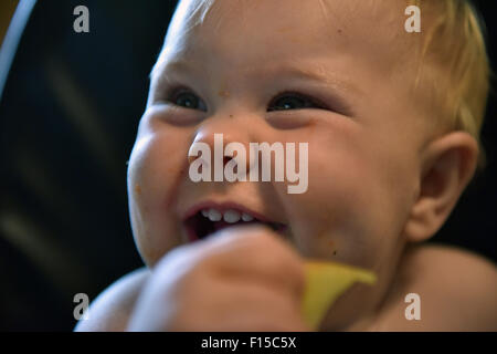 An 11 month old baby sitting in a high chair smiling with a slice of apple in her hand. Stock Photo