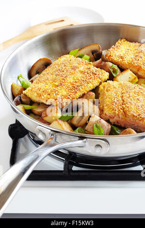 Cornmeal crusted trout fillets in a skillet with potatoes, mushrooms and green onions on a gas stove, studio shot Stock Photo