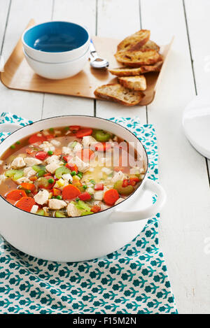 Chicken and vegetable soup in a stock pot with bread and bowls in the background, studio shot Stock Photo
