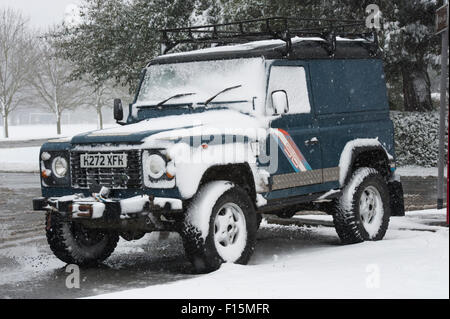 Blue Land Rover Defender 90 (rugged 4x4 vehicle) partly covered in white snow, parked at the roadside on a snowy winter day - Ilkley, England, UK. Stock Photo