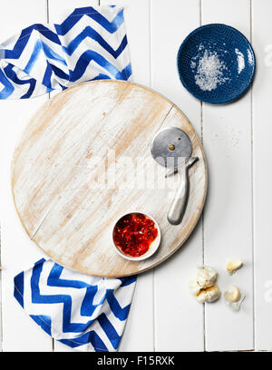 Overhead View of Wooden Cutting Board with Bowl of Chili Sauce, Pizza Cutter, Garlic Cloves, Dish with Salt, and Napkin Stock Photo
