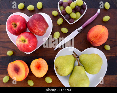 Assortment of fruit, including apples, pears, nectarines and grapes, on white heart shape plates on rustic wood table, overhead. Stock Photo