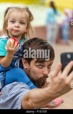 Father holds daughter piggyback and makes a cell phone self-portrait. Selfie. The concept of the modern family. Stock Photo