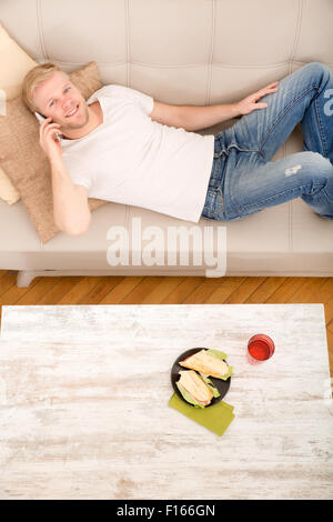 Young man with a Sandwich on the Sofa while talking on the phone. Stock Photo