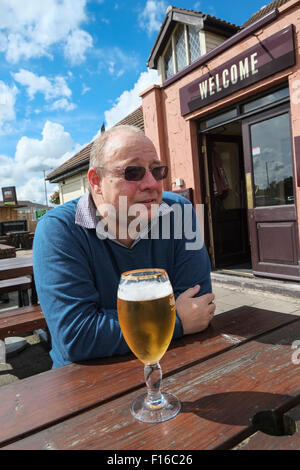 Middle-aged man sat outside a pub drinking a pint of lager with Welcome sign in background Stock Photo