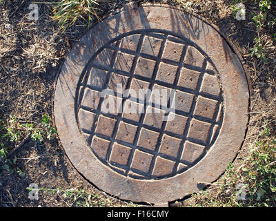 Overhead of an old and rusty manhole metal plates close-up, surrounded by soil and dry grass Stock Photo