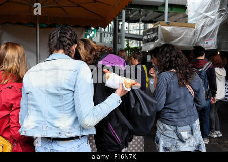 Young woman in a crowd of shoppers holding eating hotdog in bun Borough Market Southwark, South London UK  KATHY DEWITT Stock Photo