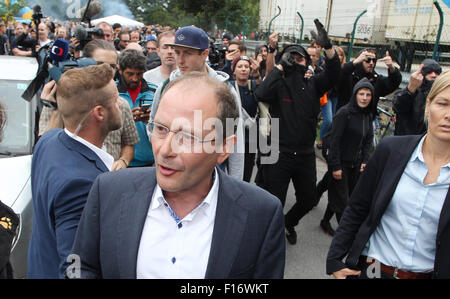 Heideau, Germany. 28th Aug, 2015. Saxony's interior minister Markus Ulbig (CDU, M) speaks to the media at a welcome party for refugees, while some of its attendees protest in the background, in Heideau, Germany, 28 August 2015. Credit:  dpa picture alliance/Alamy Live News Stock Photo