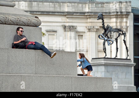 London, England, UK. Man sitting and girl climbing on the base of Nelson's Column, Trafalgar Square. 'Gift Horse' by Hans Haacke Stock Photo