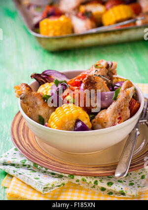 Roasted chicken with vegetables in bowl. Chicken wings, tomato, corn and onion. Green background, selective focus, rustic Stock Photo