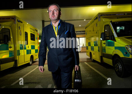 A male out-of-hours doctor, on call, holding medical bag and mobile phone, walking past a line of ambulances at a hospital UK GP Stock Photo