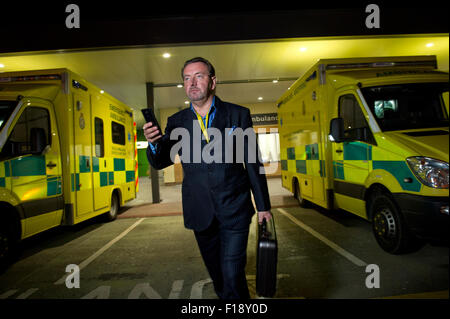 A male out-of-hours doctor, on call, holding medical bag and mobile phone, walking past a line of ambulances at a hospital UK GP Stock Photo