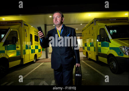 A male out-of-hours doctor, on call, holding medical bag and mobile phone, walking past a line of ambulances at a hospital UK GP Stock Photo