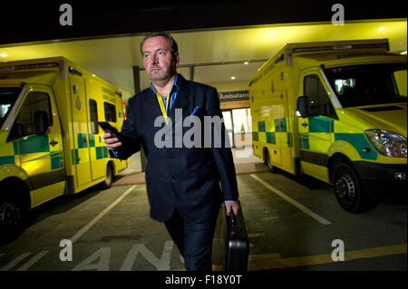 A male out-of-hours doctor, on call, holding medical bag and mobile phone, walking past a line of ambulances at a hospital UK GP Stock Photo