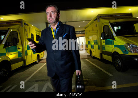 A male out-of-hours doctor, on call, holding medical bag and mobile phone, walking past a line of ambulances at a hospital UK GP Stock Photo