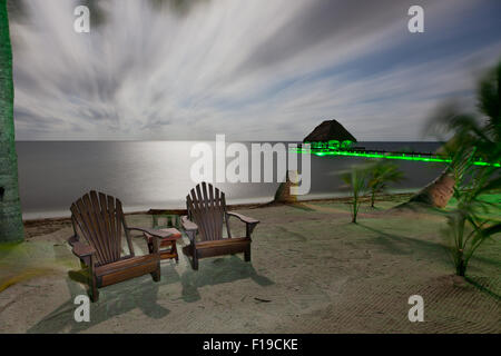 A night time beach with moonlight and green accent lights highlighting a covered dock and beach chairs in a long exposure. Stock Photo