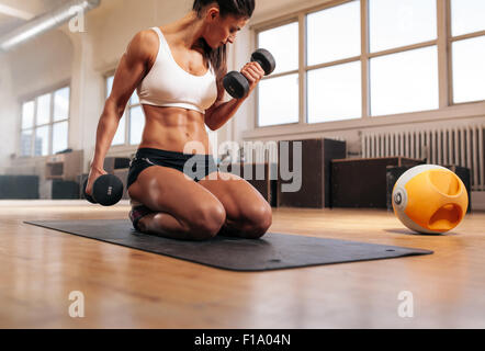 Physically fit woman at the gym lifting dumbbells to strengthen her arms and biceps. Muscular woman sitting on exercise mat look Stock Photo