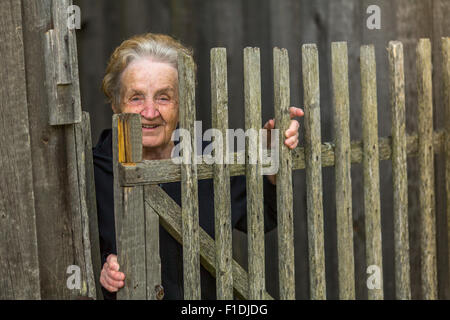 Happy old woman stands behind a wooden fence in the village. Stock Photo