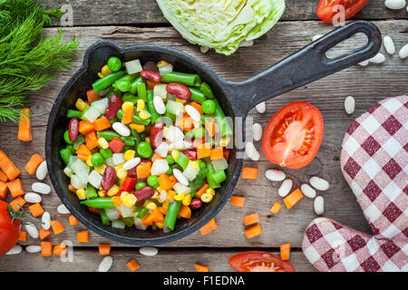Mixed vegetables in vintage frying pan, pot holder and ingredients on wooden rustic table. Top view. Stock Photo