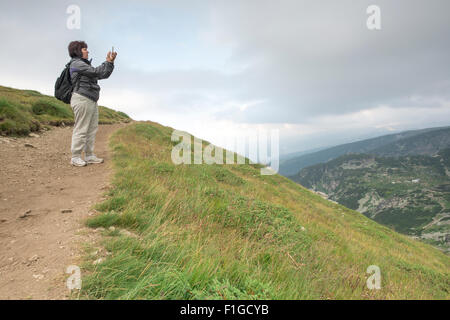 Senior woman taking photos with smartphone on the mountain. Dramatic sky Stock Photo