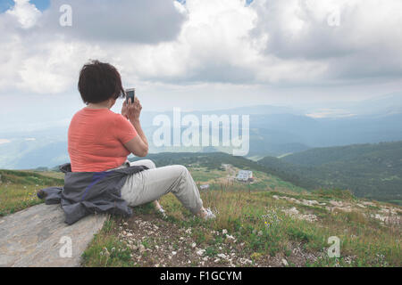 Senior woman taking photos with smartphone on the mountain. Dramatic sky Stock Photo