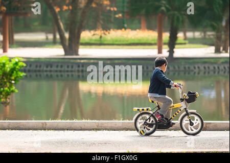 Child playing alone, view of a child cycling past a lake in Hanoi's Botanical Garden, Vietnam. Stock Photo