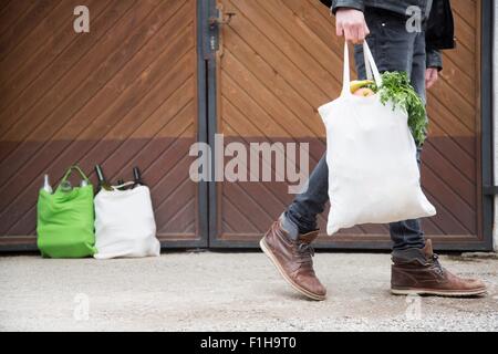 Teenage boy carrying reusable shopping bag full of fruit and veg, with bottles for recycling in yard Stock Photo