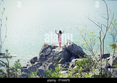 Mid adult woman, standing on rock beside lake, in yoga position, rear view Stock Photo