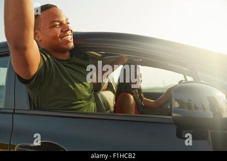 Young man leaning out of car window smiling, arms raised Stock Photo