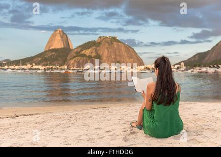 Mid adult woman sitting in beach, reading book, Sugarloaf mountain in background, Botafogo bay, Rio de Janeiro, Brazil Stock Photo