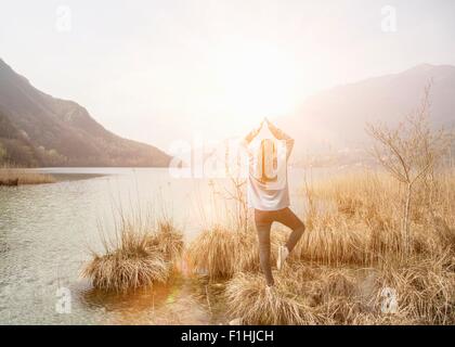 Rear view of young woman practicing yoga on lakeside, Lake Mergozzo, Verbania, Piemonte, Italy Stock Photo