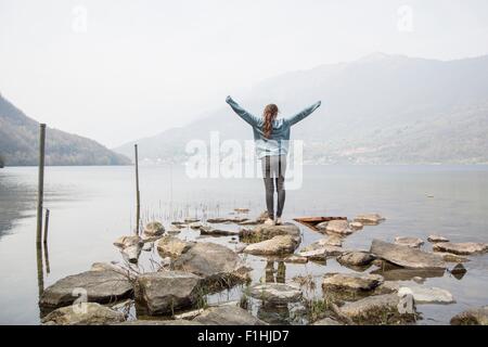 Rear view of young woman on rocks with arms raised at Lake Mergozzo, Verbania, Piemonte, Italy Stock Photo