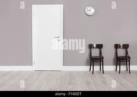 Two wooden chairs in a waiting room with a clock hanging on the wall above them Stock Photo