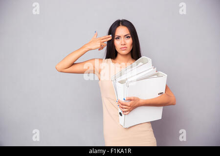 Businesswoman holding folders and making gun gesture to her head over gray background Stock Photo