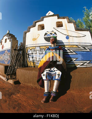 Ndebele woman in traditional dress leaning against wall of brightly coloured village. Botshabelo Mpumalanga South Africa Africa Stock Photo