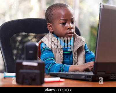 Young African boy sitting at his father's desk, playing on the laptop Stock Photo