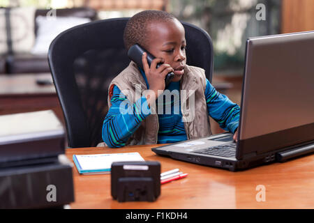 Young African boy sitting at his father's desk, playing on the laptop and talking on telephone Stock Photo