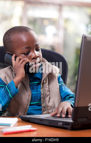 Young African boy sitting at his father's desk, playing on the laptop and talking on telephone Stock Photo